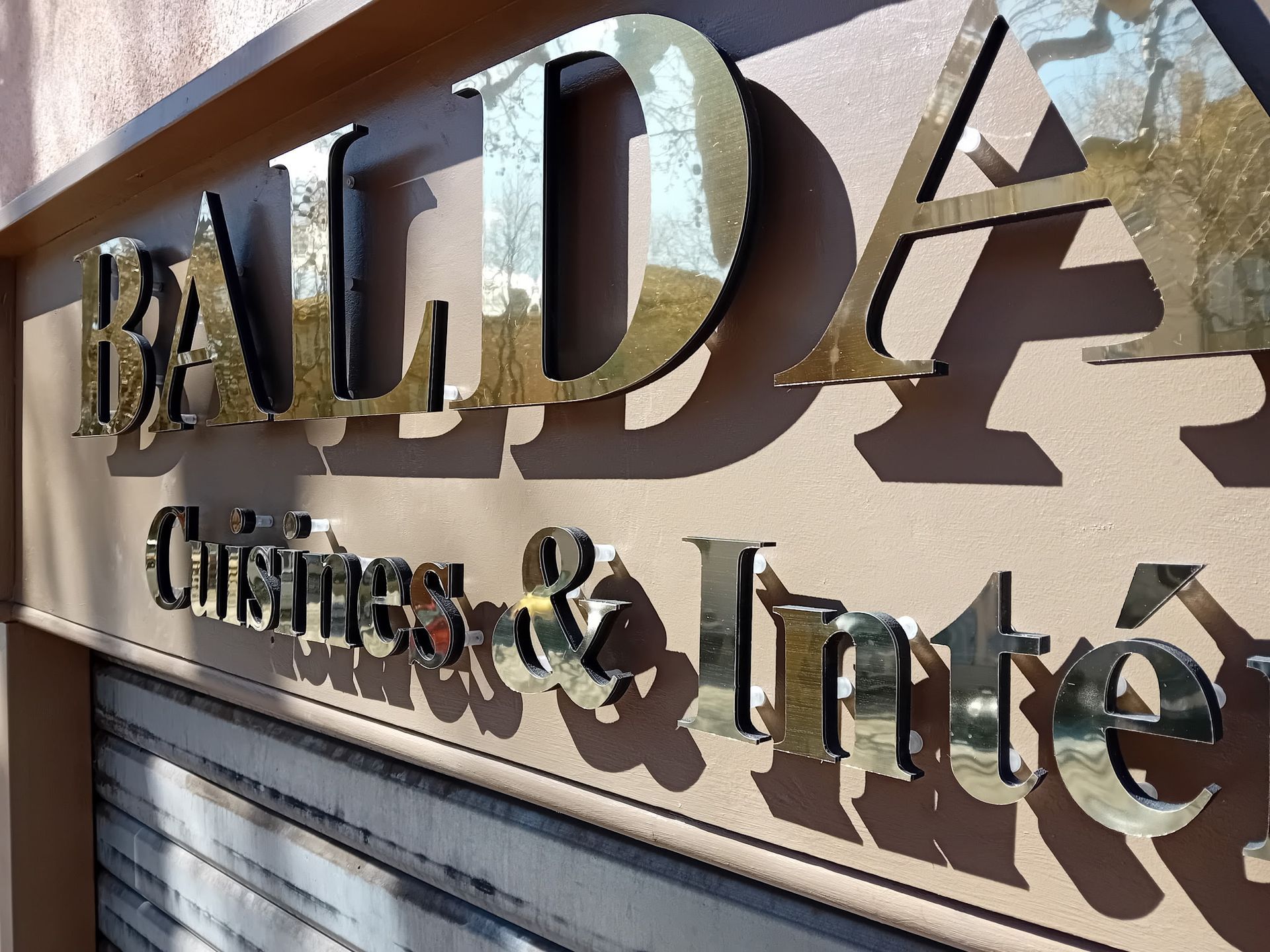 Une enseigne métallique portant l'inscription Baldano Cuisines & Intérieurs est fixée sur un mur beige à l'extérieur d'un bâtiment.