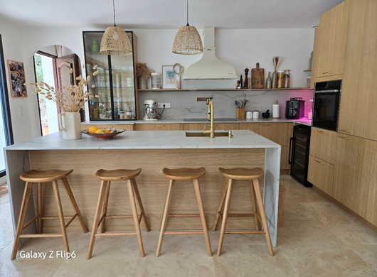 Cuisine moderne et lumineuse avec îlot central, quatre tabourets en bois, suspensions lumineuses et meubles de cuisine clairs.