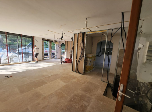 Salon partiellement rénové avec murs apparents, grandes fenêtres et matériaux de construction au sol