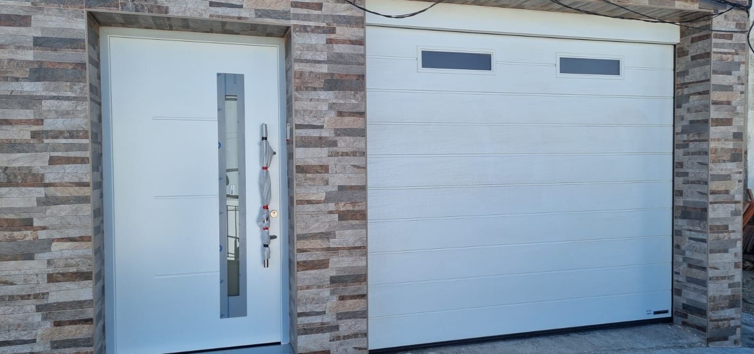 Una puerta exterior blanca con una ventana vertical y una amplia puerta de garaje blanca con paneles horizontales, integrada en un muro de piedra.