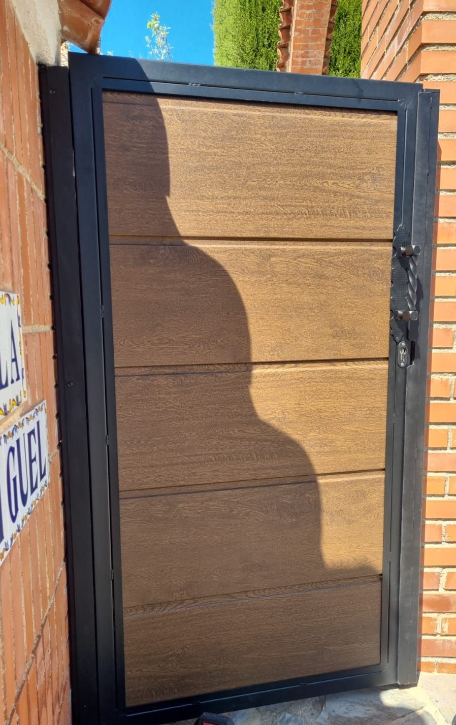 Una puerta con marco negro y paneles horizontales con textura de madera, incrustada en una pared de ladrillo, con la sombra de una persona proyectada en la parte frontal.