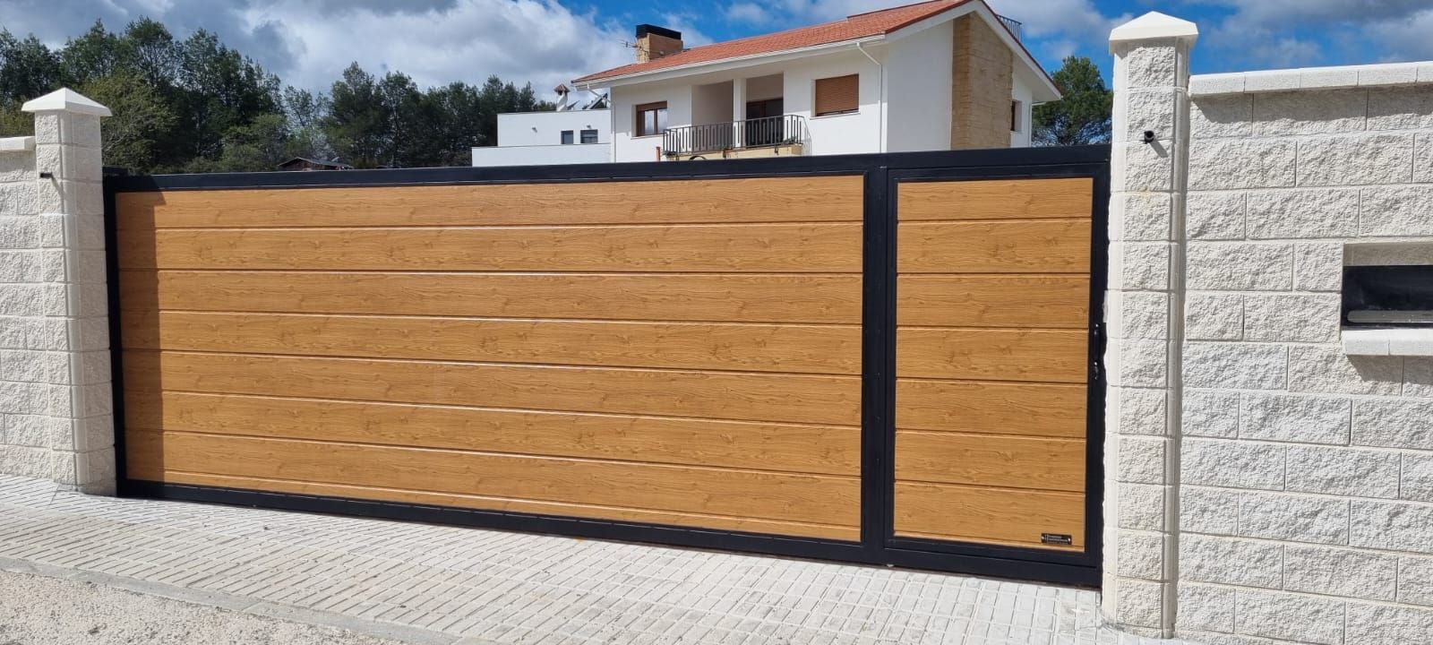 Una amplia puerta horizontal con paneles que imitan la veta de la madera y un marco negro, situada entre columnas de piedra frente a una casa blanca.
