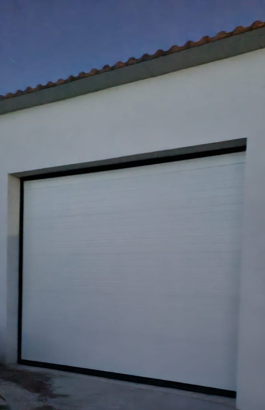 Una puerta de garaje seccional blanca integrada en una pared lisa con un borde de techo oscuro contra un cielo despejado.