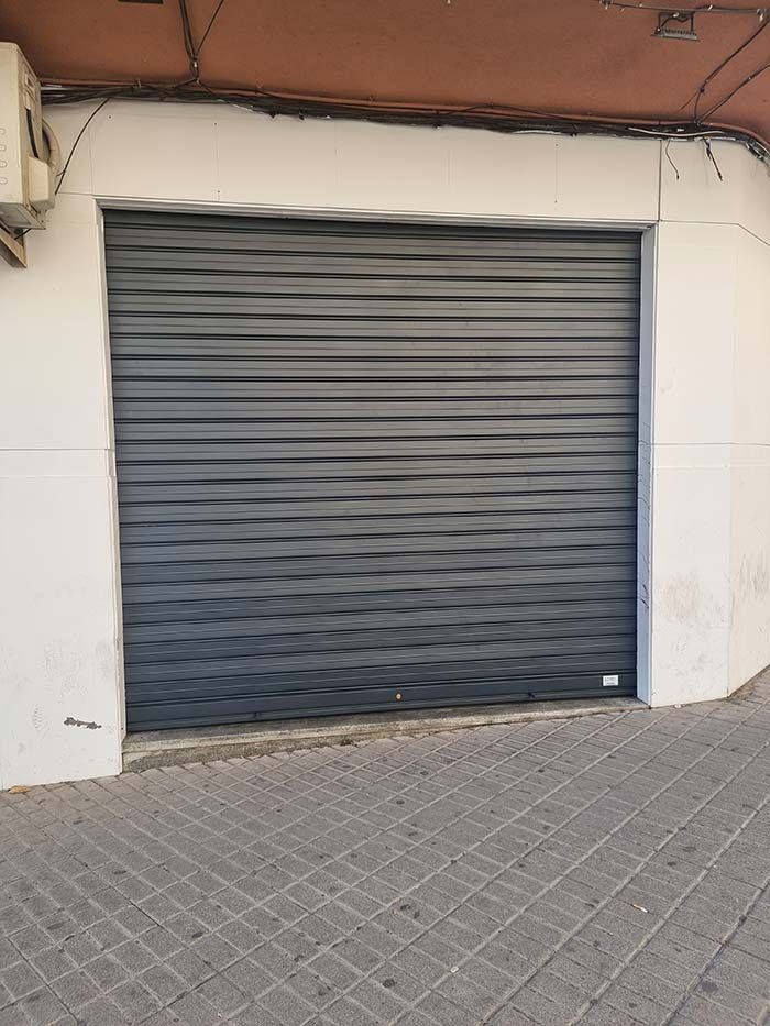 Puerta de seguridad metálica gris, cerrada, en un edificio de paredes blancas. Acera al frente.