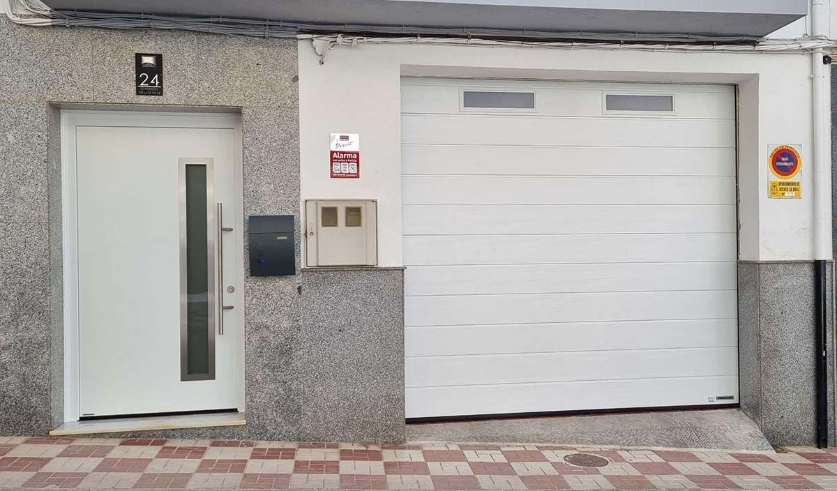 Puerta blanca y puerta de garaje contiguas. Exterior del edificio con fachada de granito.