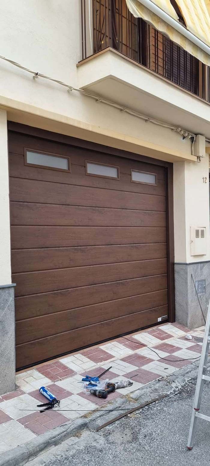 Puerta de garaje marrón con tres pequeñas ventanas encima, herramientas en la acera a cuadros.
