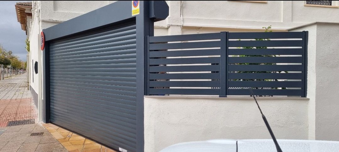 Puerta de garaje gris oscuro junto a una valla de listones horizontales a juego. La antena de un coche sobresale de la parte inferior.