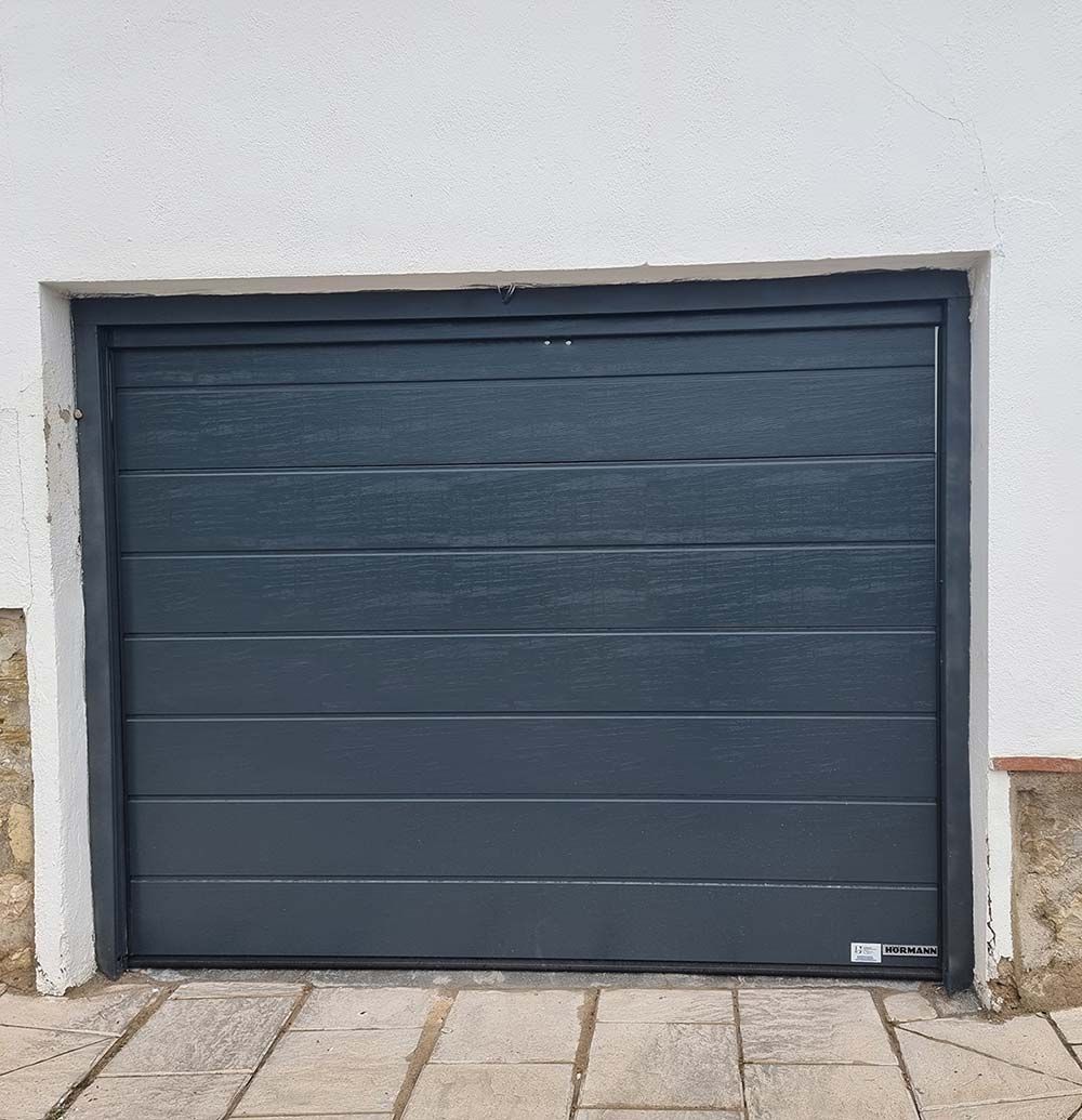 Puerta de garaje de panel horizontal de color gris oscuro en un edificio de paredes blancas con acera de piedra.