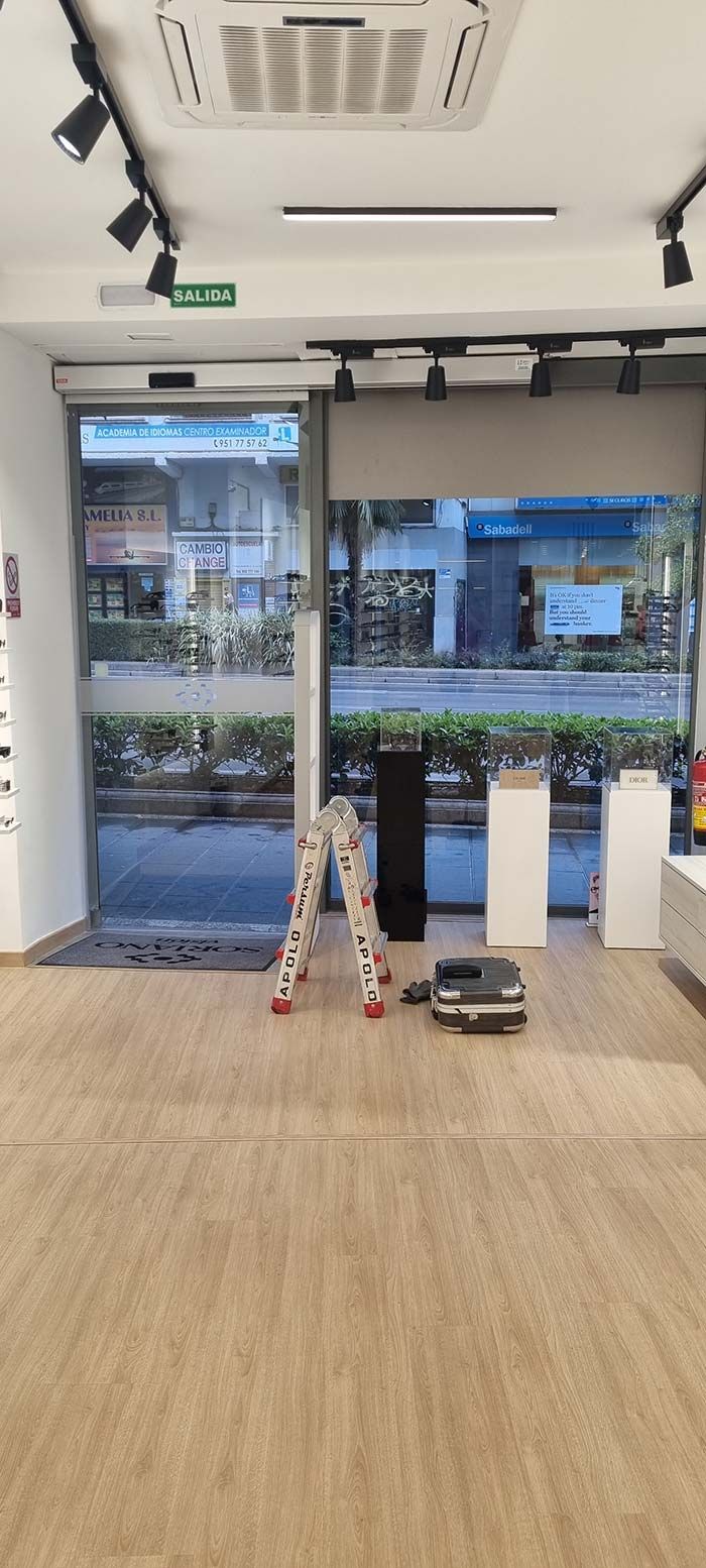 Vista interior de la tienda; escalera cerca de la entrada. Puertas de cristal, con la calle visible a través de ellas. Columnas blancas y equipo en el suelo.