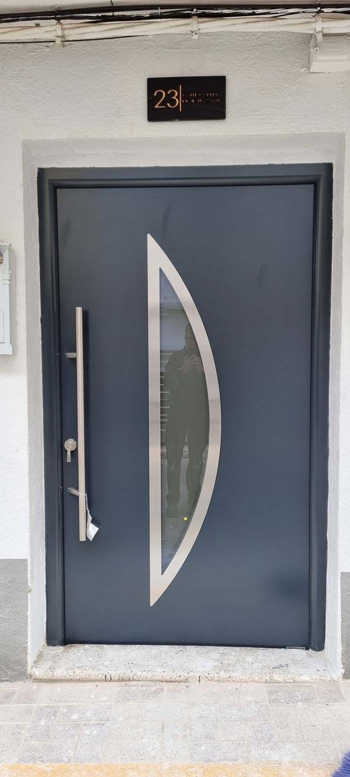 Puerta gris con ventana en forma de media luna. Tiene un tirador largo y una placa con el número