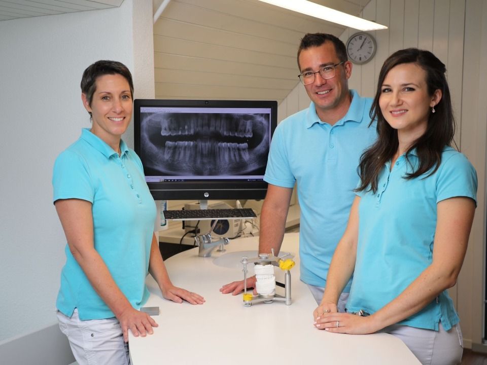 Drei Personen in blauen Poloshirts stehen in einer Klinik hinter einem zahnärztlichen Untersuchungstisch mit einem Röntgenmonitor.