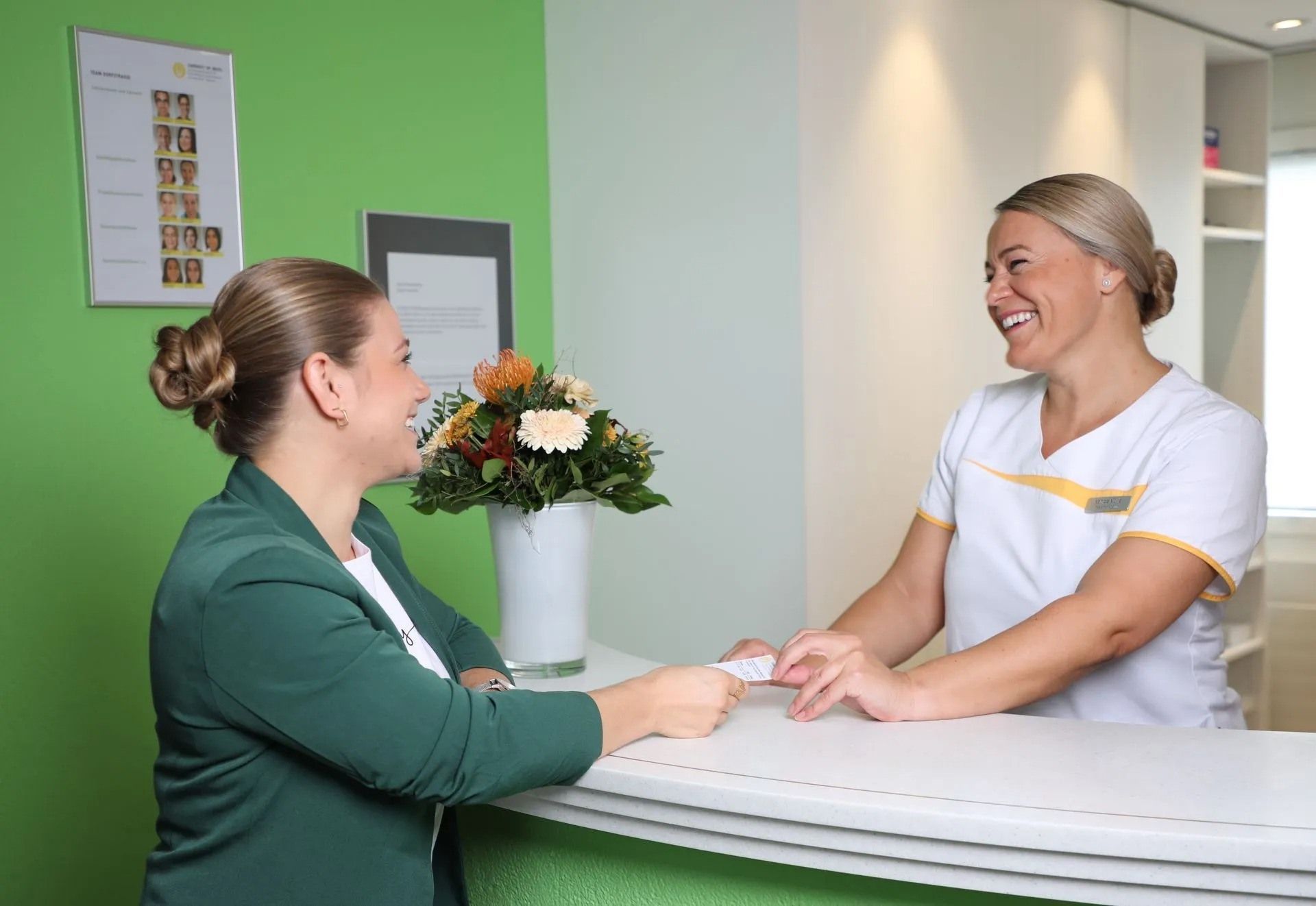 Eine Rezeptionistin begrüsst lächelnd und per Handschlag einen Kunden am Empfang einer Klinik; im Hintergrund sind Blumen zu sehen.