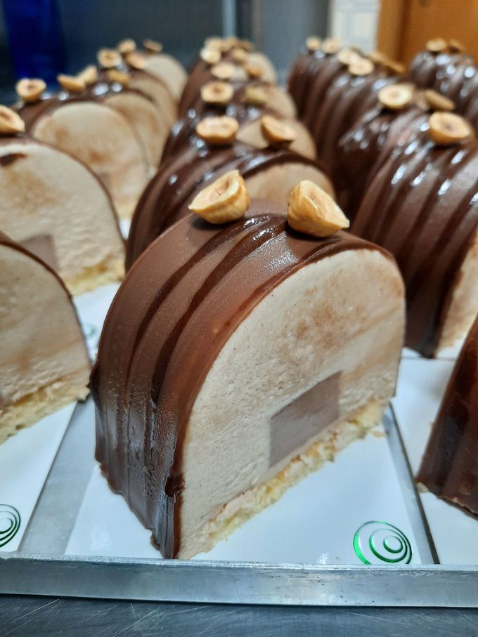 Filas de pasteles glaseados con chocolate y cubiertos de frutos secos, expuestos en bandejas en una panadería.