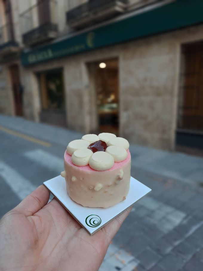 En una calle de la ciudad, una mano sostiene un pequeño postre de crema decorado con flores rosas y blancas.