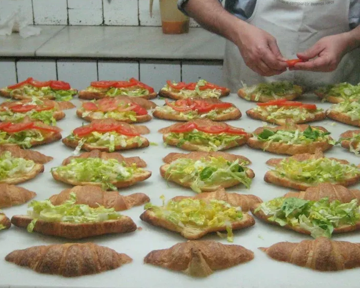 Un trabajador de la cocina está preparando hileras de croissants cubiertos con lechuga, tomate y queso.