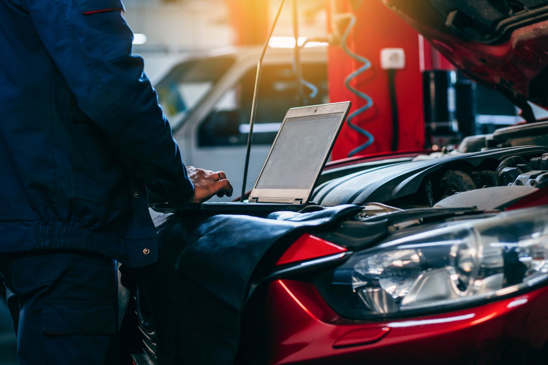 Un mécanicien en uniforme bleu utilise un ordinateur portable pour diagnostiquer une voiture rouge, capot ouvert, dans un garage.