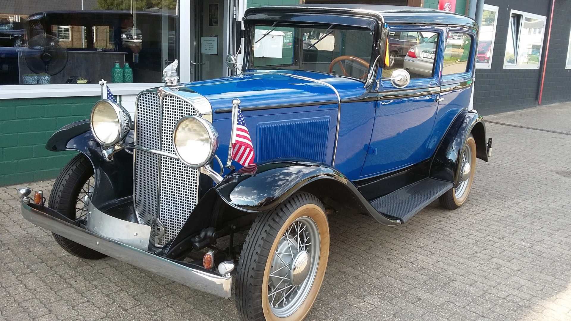 Ein altes blaues Auto mit einer amerikanischen Flagge auf der Vorderseite ist vor einem Gebäude geparkt.