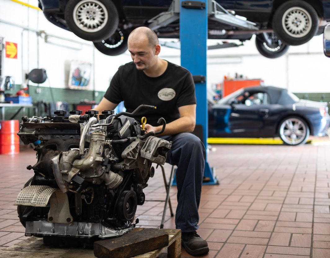 Ein Mechaniker arbeitet in einer Autowerkstatt an einem Automotor; im Hintergrund sind hochgelegte Autos zu sehen.