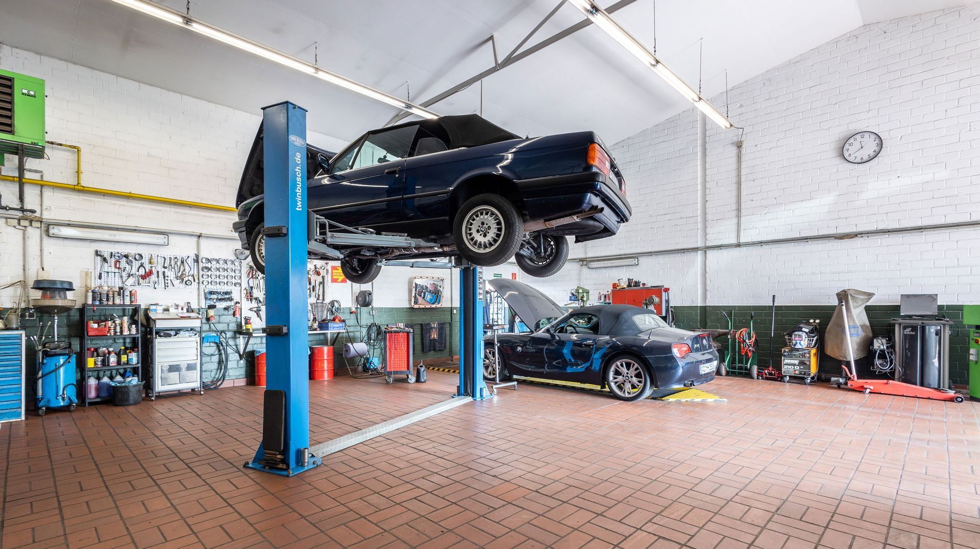 Autowerkstatt mit einem Wagen, der auf einer Hebebühne angehoben ist, und einem darunter Auto in einer hellen Werkstatt.