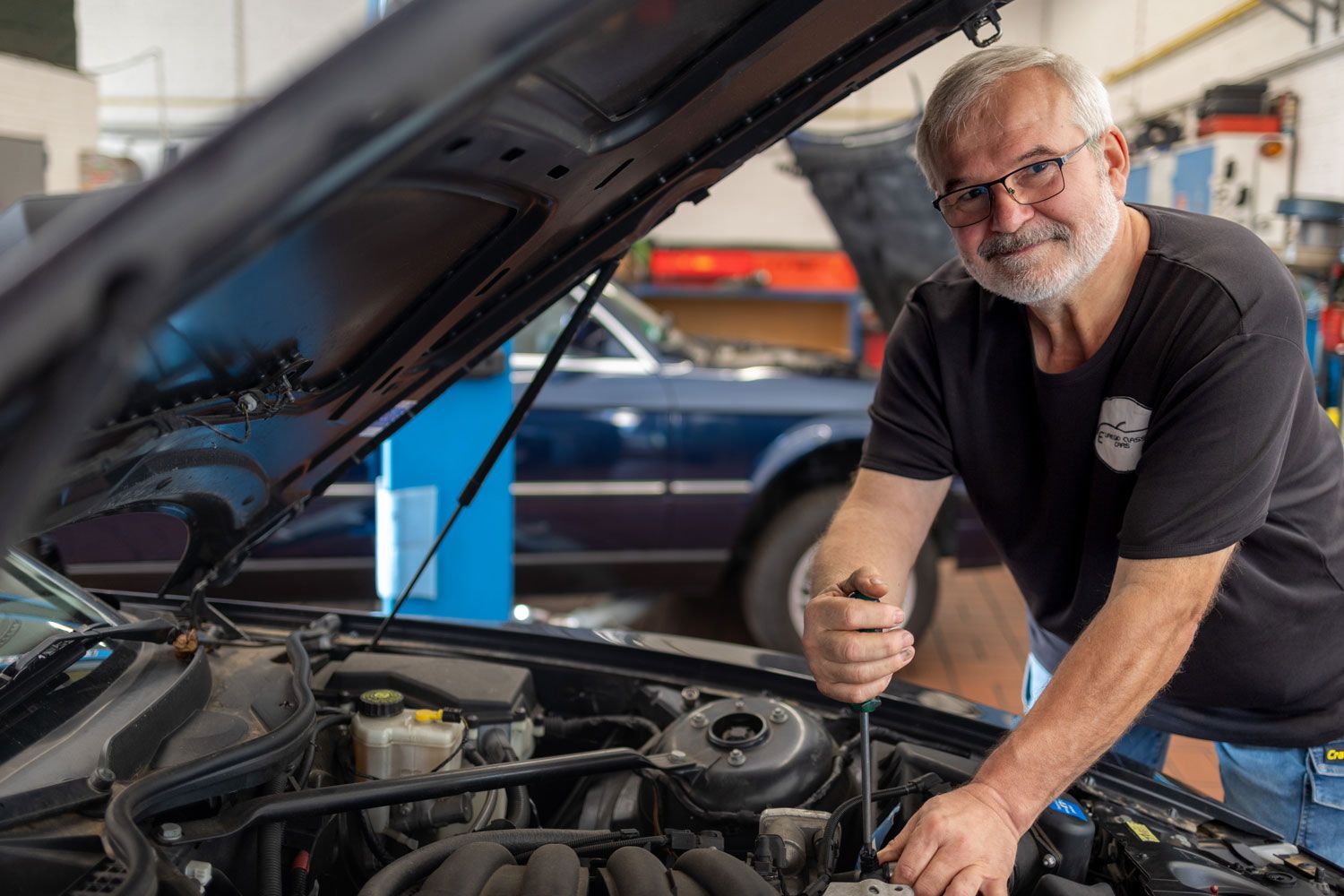 Ein Mechaniker beugt sich in einer Werkstatt über die offene Motorhaube eines Autos und benutzt ein Werkzeug am Motor.