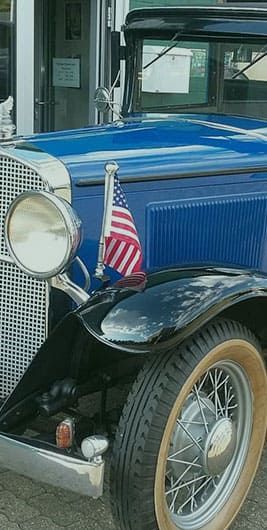 Ein altes blaues Auto mit einer amerikanischen Flagge auf der Motorhaube steht vor einem Gebäude.