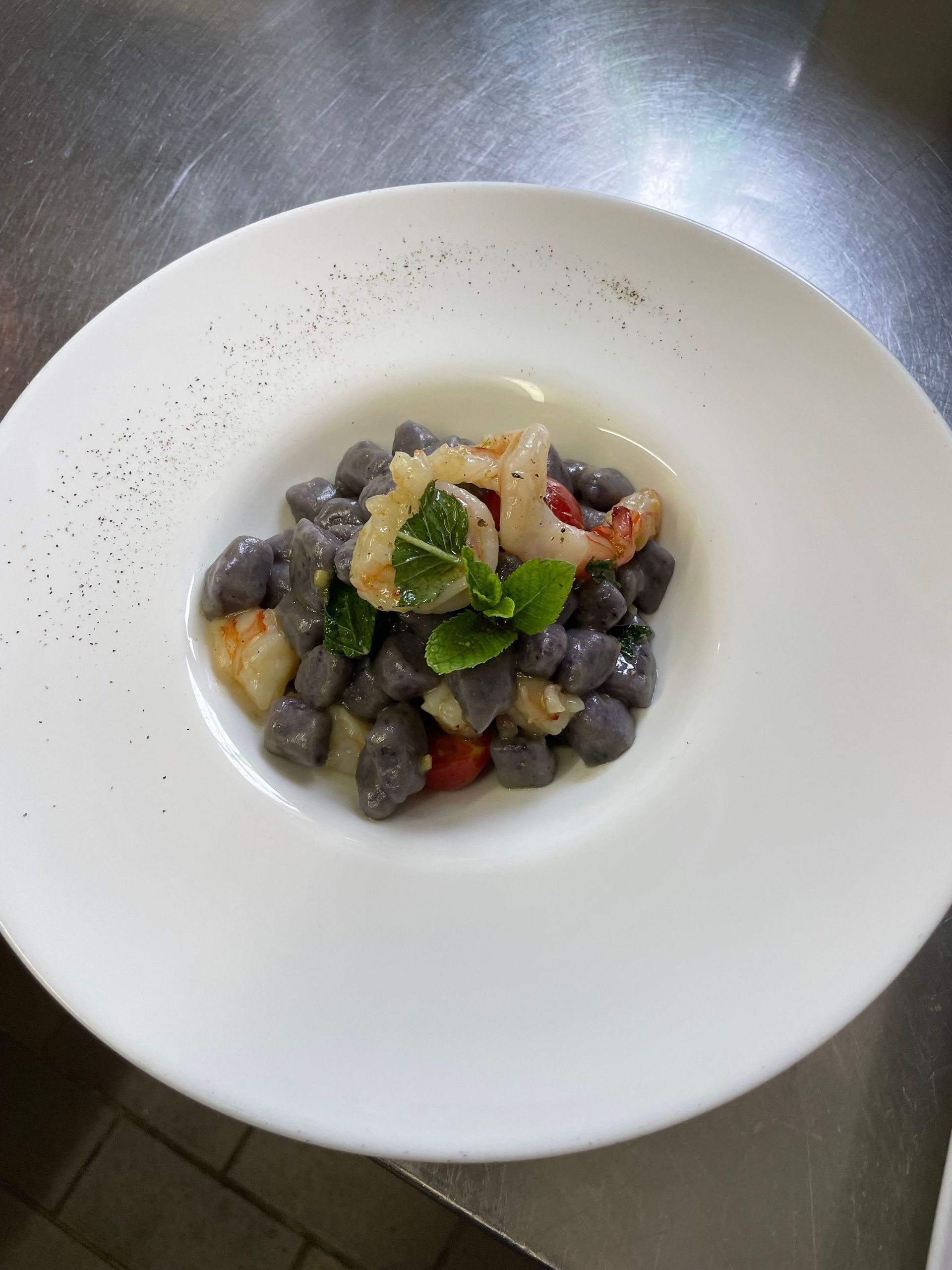 Ein weißer Teller mit dunklen Gnocchi, Garnelen, Tomaten und Basilikum.