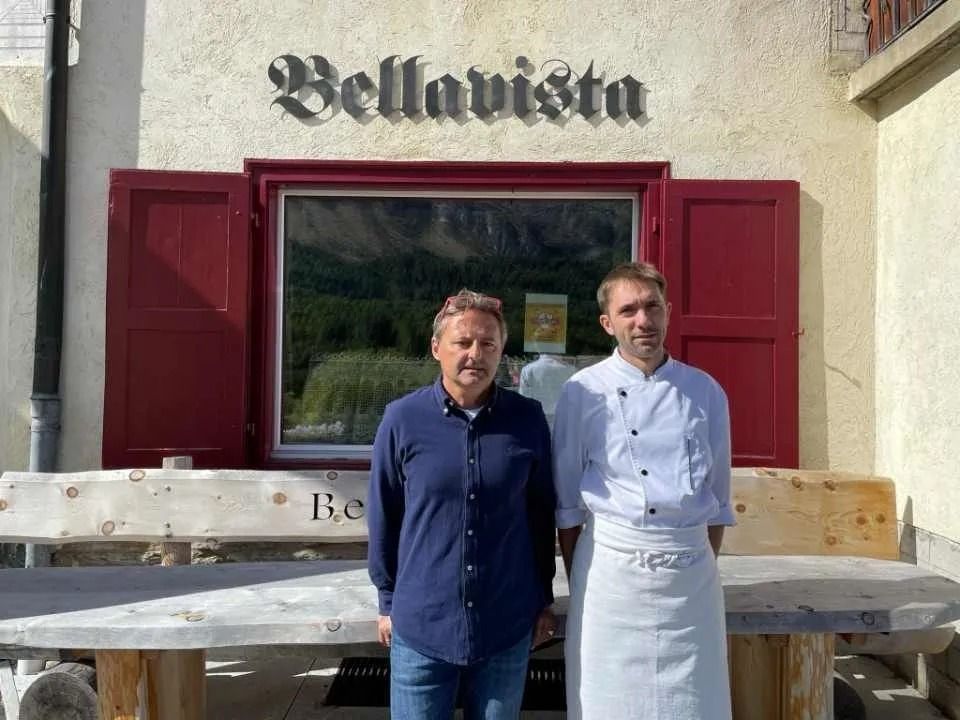 Zwei Männer stehen vor dem Restaurant „Bellavista“: einer im blauen Hemd, der andere in der weißen Kochkleidung. Rote Fensterläden und eine Holzbank.
