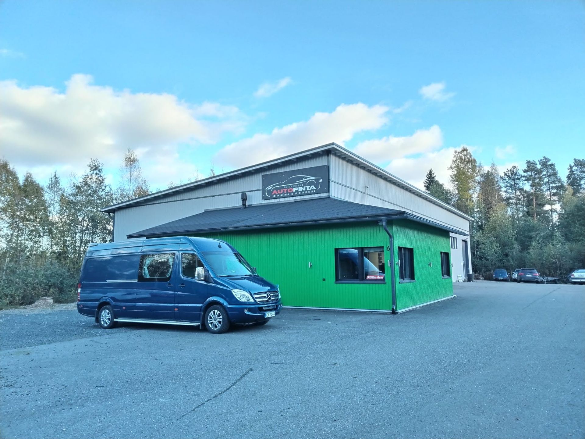 Sininen pakettiauto pysäköitynä kirkkaanvihreän rakennuksen viereen pilvisen taivaan alle.