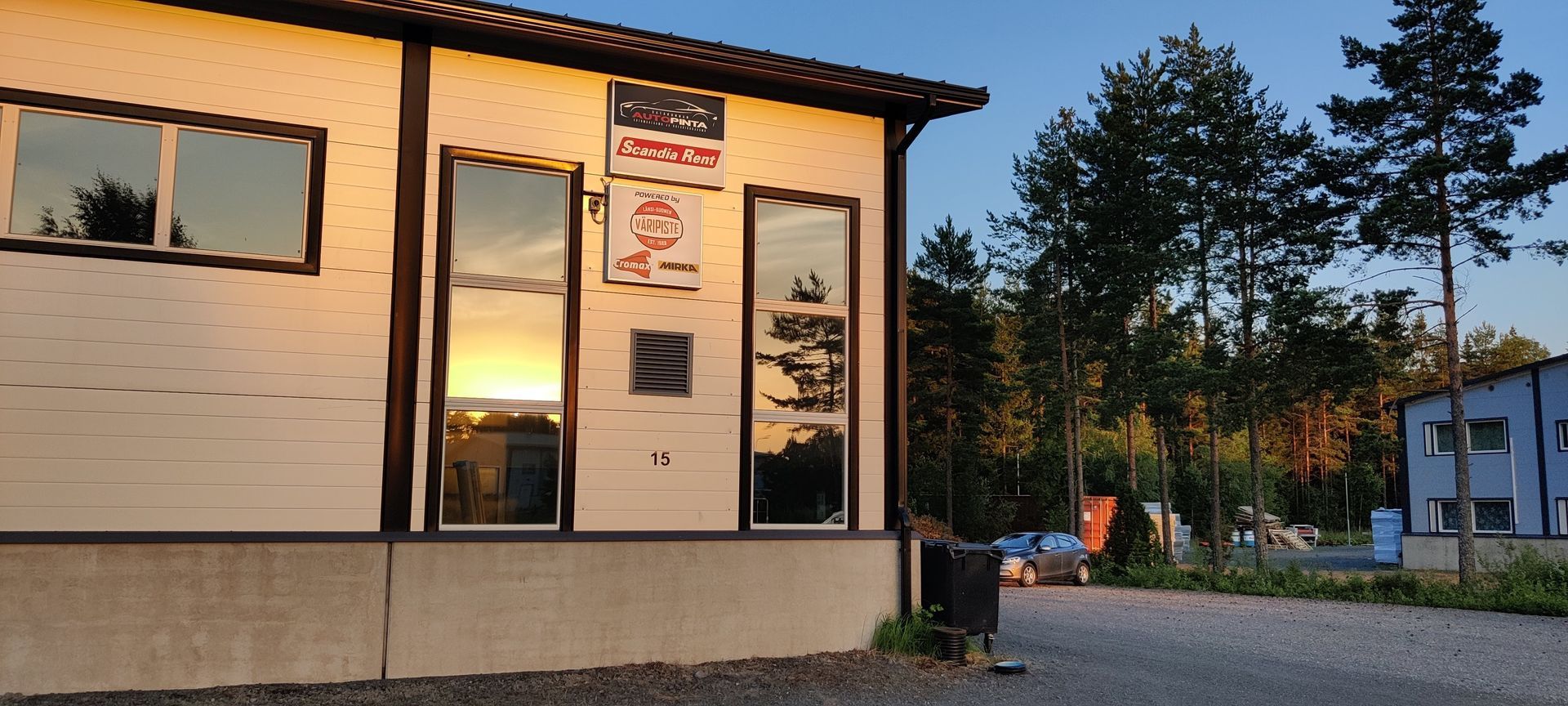Pieni valkoinen rakennus auringonlaskun aikaan heijastavilla ikkunoilla, kyltti oven lähellä ja puita ja soratontti lähistöllä