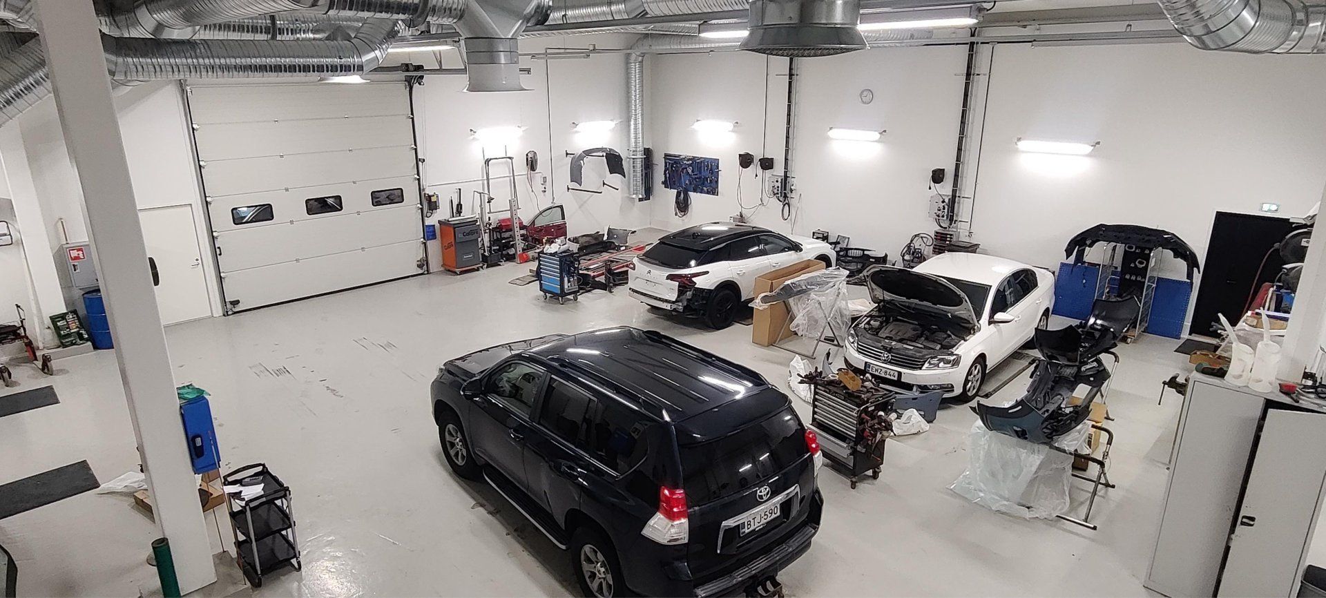 Laajakuvanäkymä kiireisestä autokorjaamosta, jonka sisällä työskentelee useita autoja ja mekaanikkoja