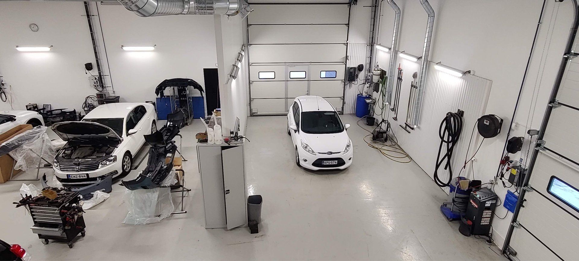 Leveä autotallityöpaja, jonka keskellä on valkoinen auto ja sivuilla työkaluja, laitteita ja työpisteitä