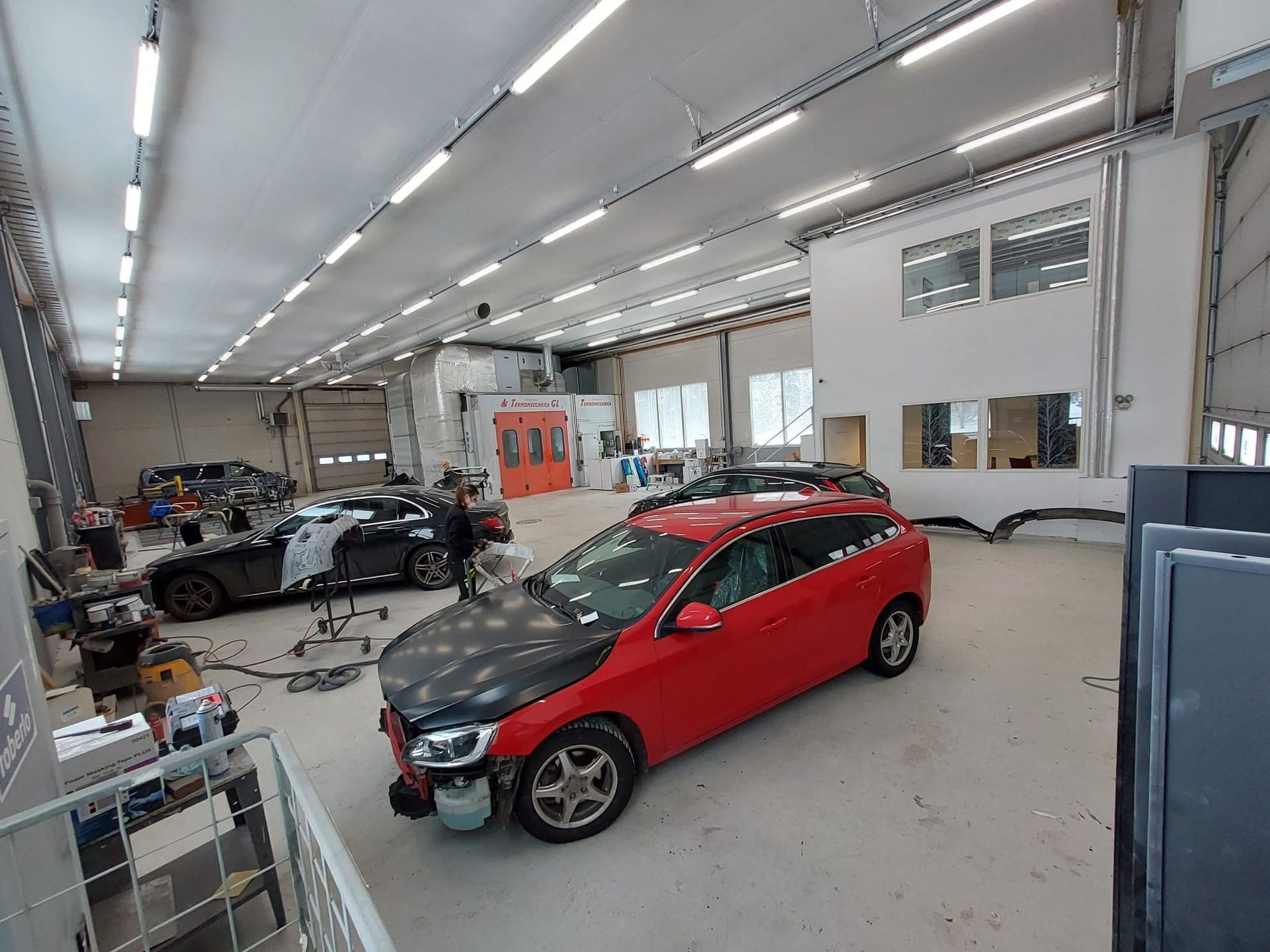 Punainen viistoperä kirkkaassa autokorjaamossa, jossa on työkaluja, hyllyjä ja musta auto taustalla