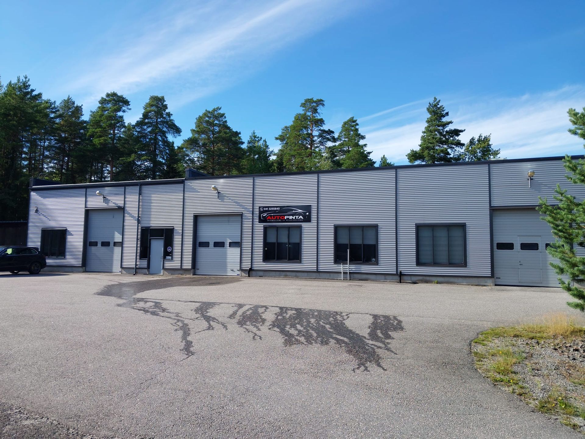 Yksikerroksinen metallinen teollisuusrakennus, jossa on useita autotallin ovia sinisen taivaan alla, edessä soraa.