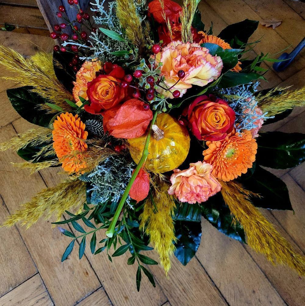 Ein Herbststrauß mit orangefarbenen Rosen, Gerbera-Gänseblümchen, einem kleinen gelben Kürbis und getrockneten Ziergräsern.