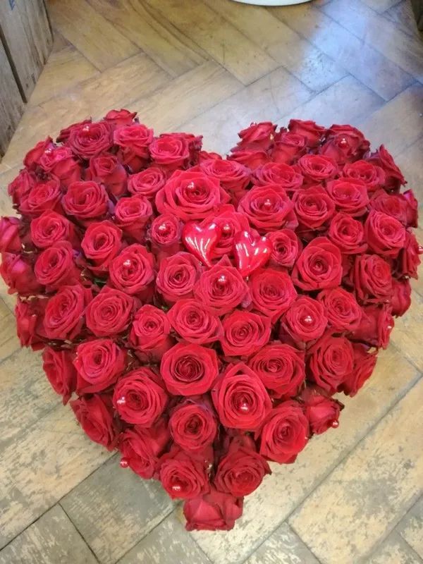 Ein herzförmiges Arrangement aus zahlreichen leuchtend roten Rosen ruht auf einem hellen Holzboden.