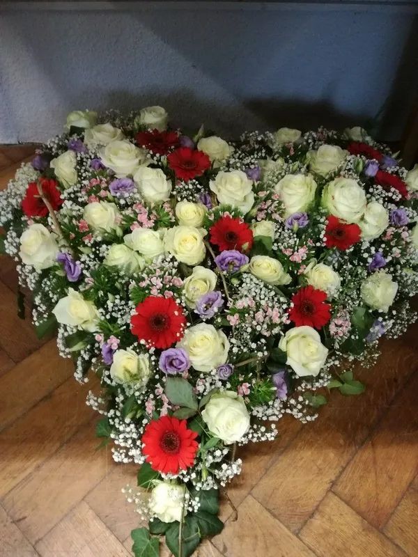 Ein herzförmiges Blumenarrangement mit roten Gerbera-Gänseblümchen, weißen Rosen, violetten Akzentblumen und Schleierkraut.