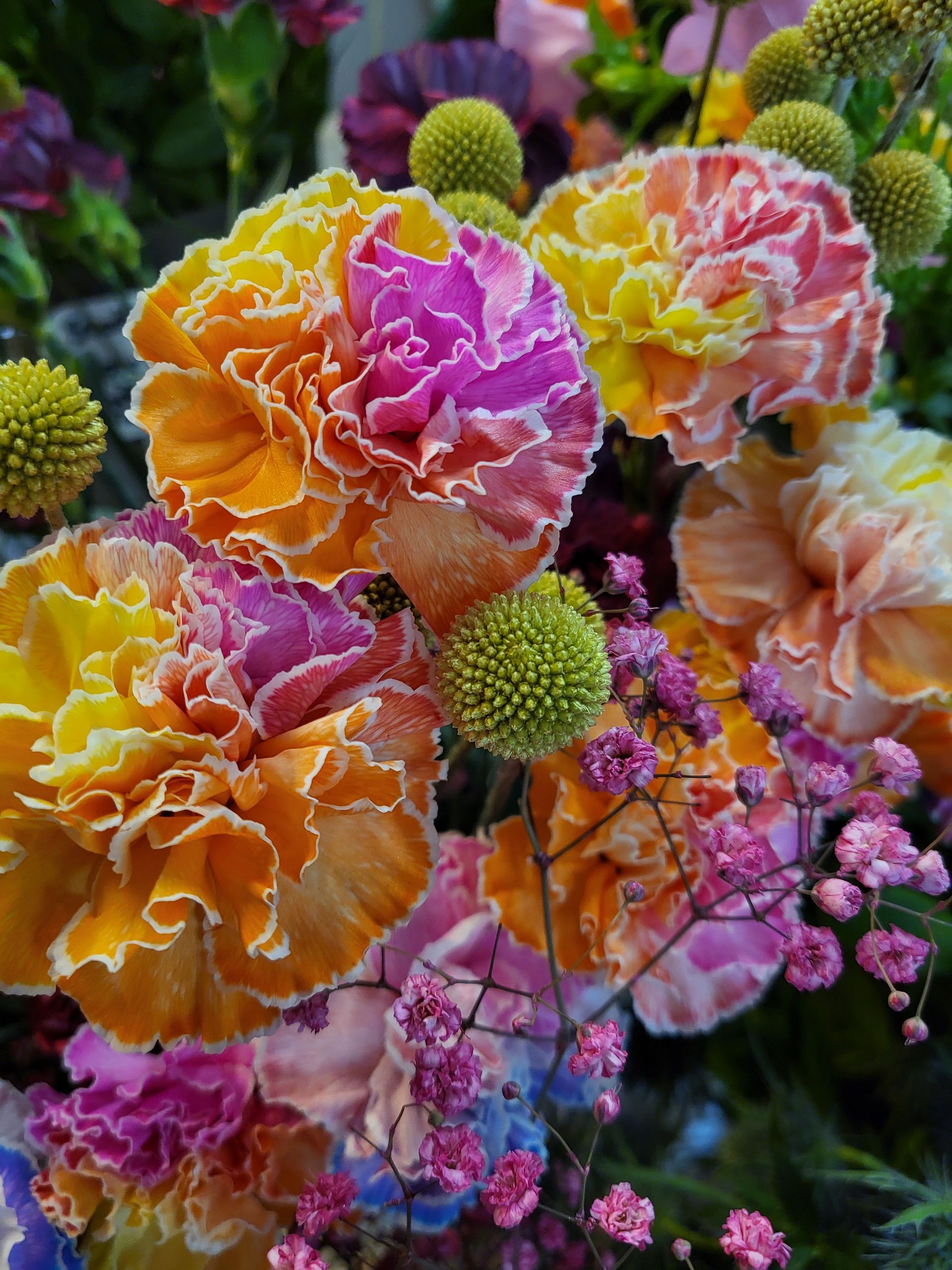 Ein Strauß aus leuchtenden, gerüschten orangefarbenen und violetten Nelken mit kleinen gelben Kugelblumen und rosa Blüten.
