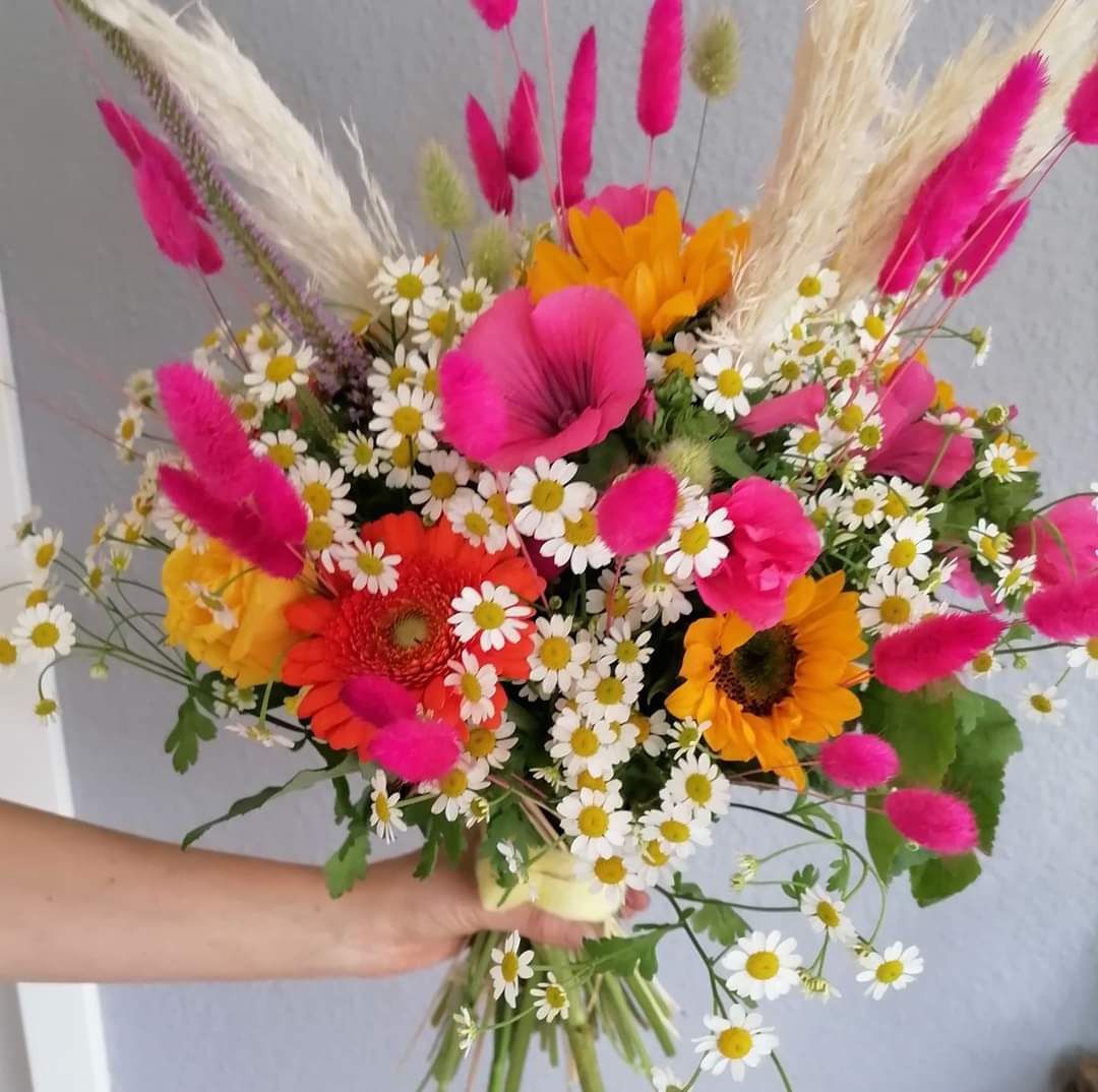 Eine Hand hält einen farbenfrohen Blumenstrauß mit Sonnenblumen, Gänseblümchen und leuchtend pinkfarbenen Trockengräsern.