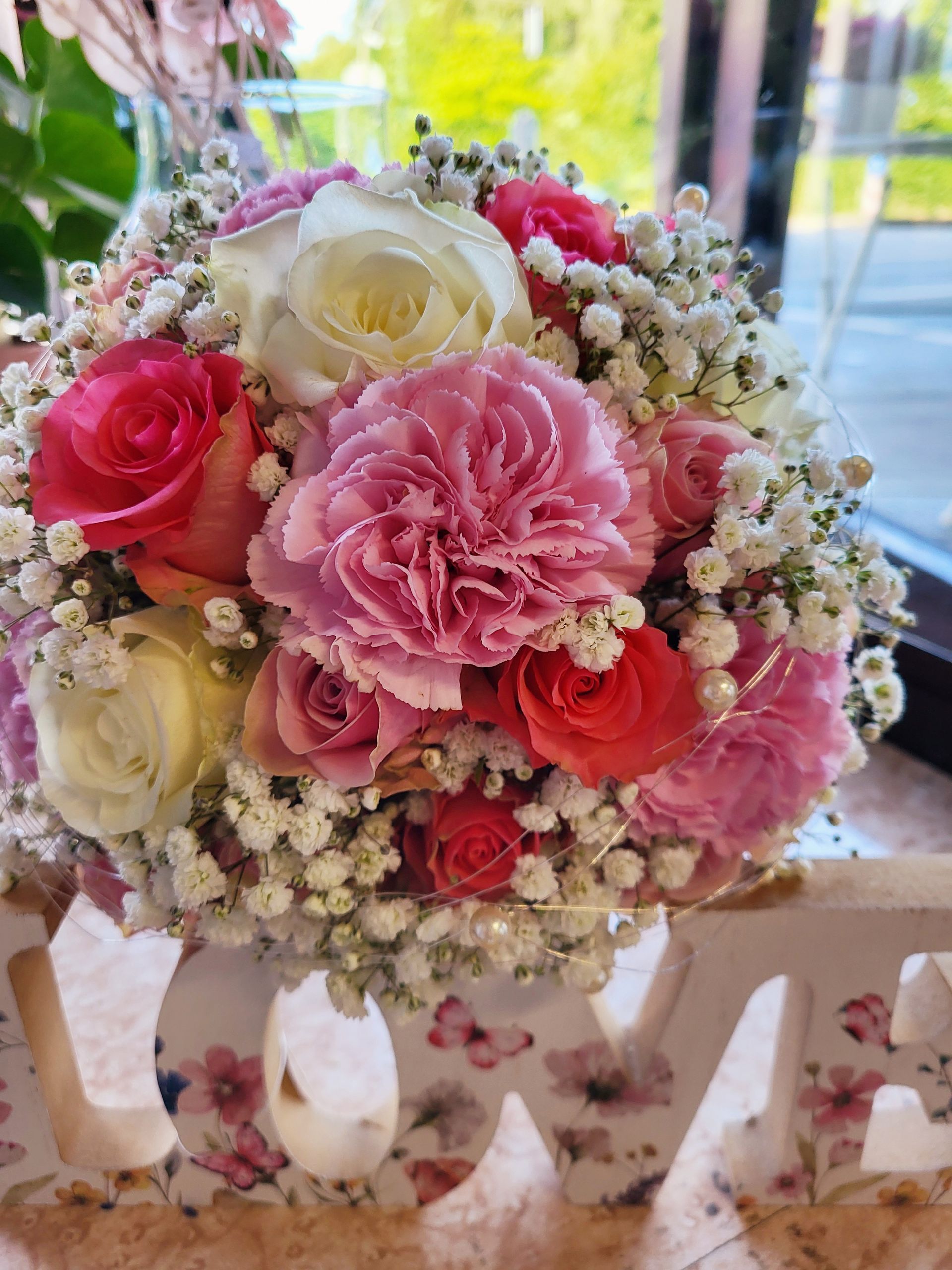 Ein Strauß aus rosa und weißen Rosen, Nelken und Schleierkraut thront auf einem hölzernen „LOVE“-Schild.