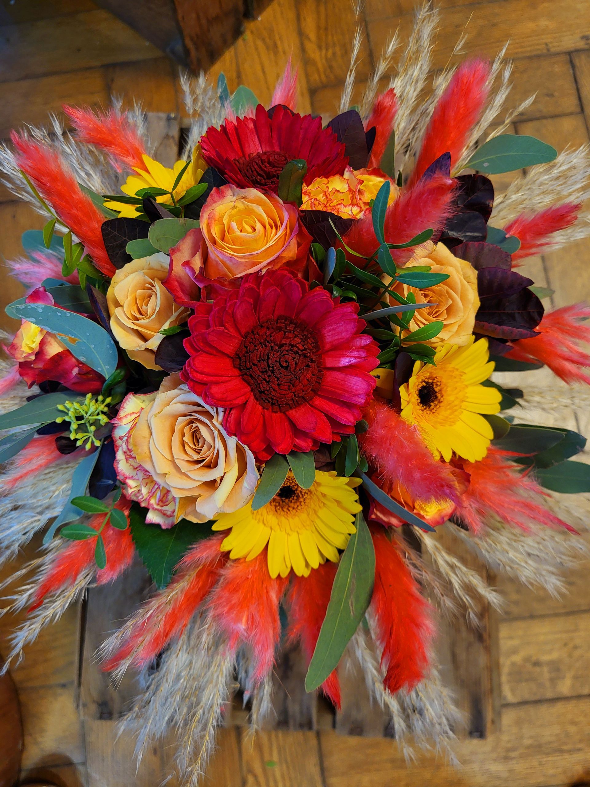 Ein leuchtender Herbststrauß mit roten und gelben Gerbera-Gänseblümchen, orangefarbenen Rosen und farbenfrohen Ziergräsern.