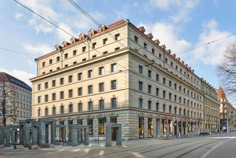 Grosses beiges Gebäude mit rotem Dach in einer Stadtstrasse; Skulpturen davor. Sonniger Tag, blauer Himmel.