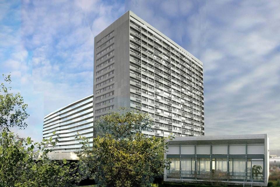 Hohe Krankenhausgebäude aus Beton mit mehreren Ebenen, Bäume im Vordergrund und ein kleines rechteckiges Gebäude.