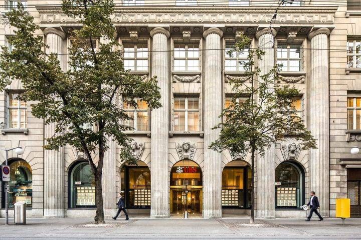 Elegantes Gebäude mit Säulen und Bögen, Schaufenster, zwei Personen gehen vorbei.