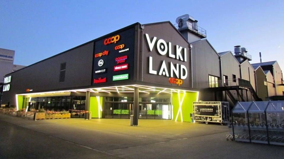 Die Aussenfassade des Einkaufszentrums „Volki Land“ ist in der Abenddämmerung mit neongrünen Akzenten beleuchtet; verschiedene Ladenschilder sind zu sehen.