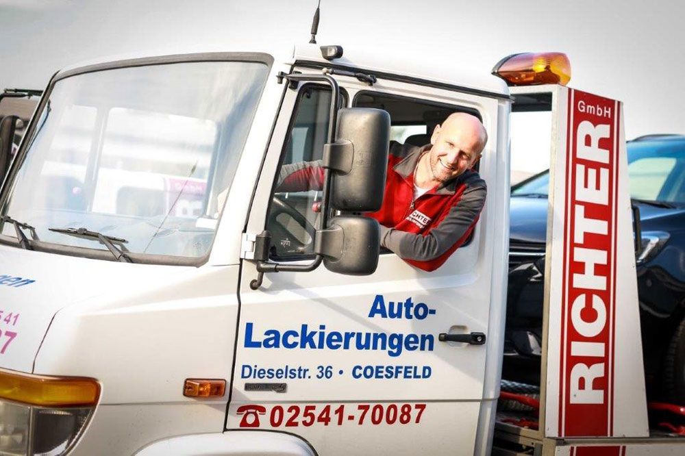 Ein Mann lehnt sich aus einem Abschleppwagen. Der Wagen ist weiß mit roter und blauer Aufschrift. Der Mann lächelt.