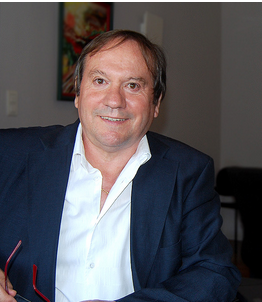 Un homme en blazer bleu foncé et chemise blanche à col ouvert sourit à l'intérieur.