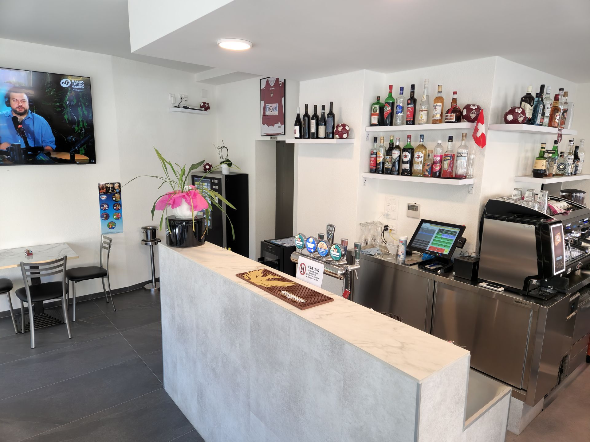 Interno di un bar: pareti bianche, bancone, scaffali con bottiglie, schermo TV e zona salotto.