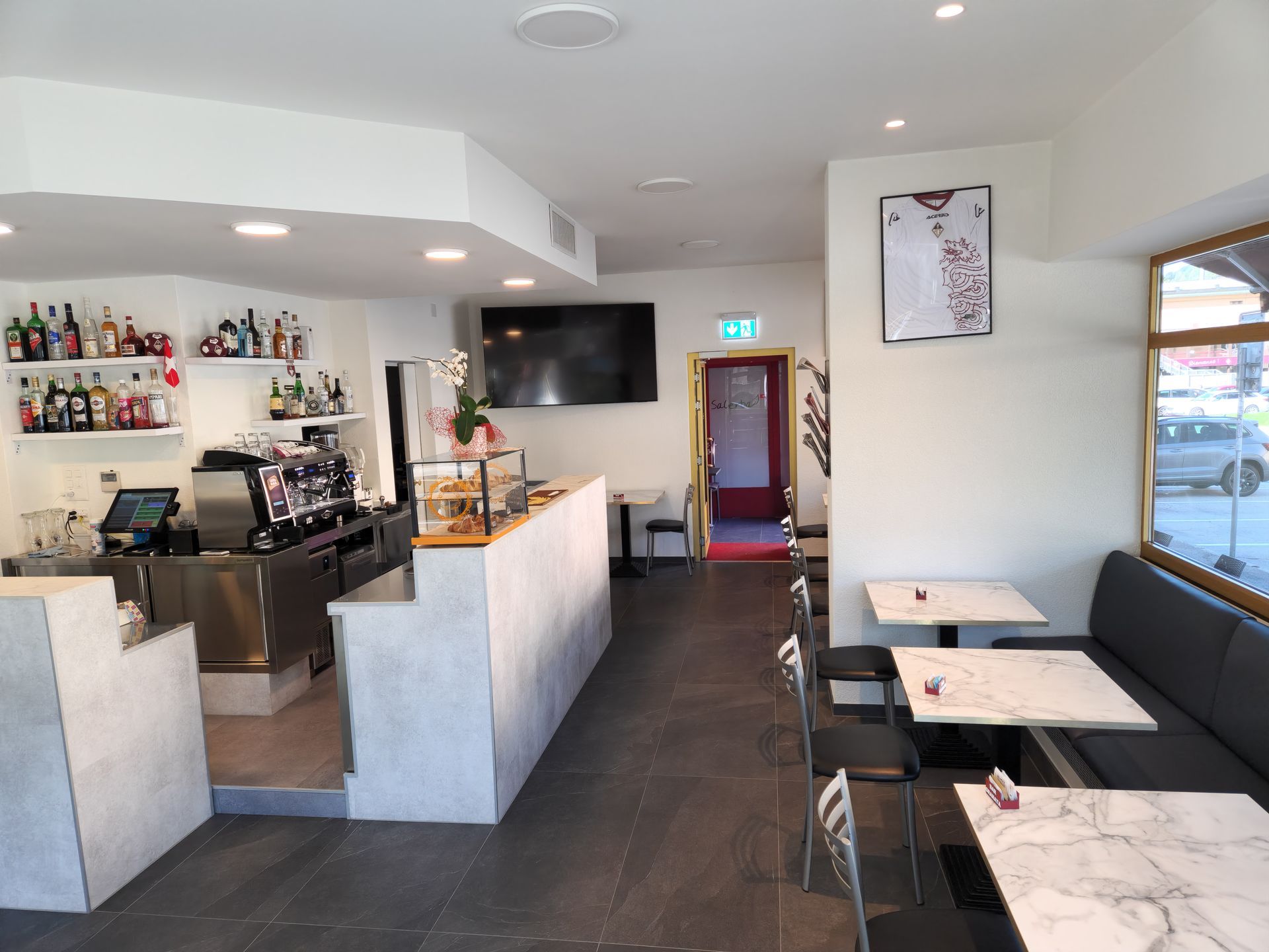 Interno di un bar con bancone, tavoli e un ingresso che conduce a una sala sul retro.