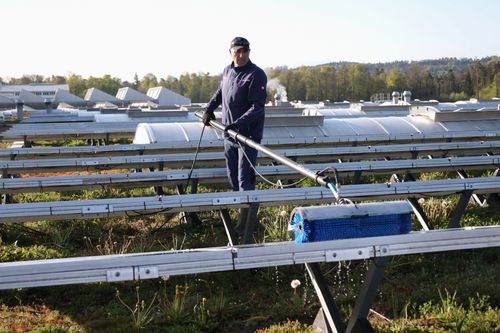 Eine Person in orangefarbener Arbeitskleidung reinigt mit einem Abzieher Solarpaneele auf einem Dach.