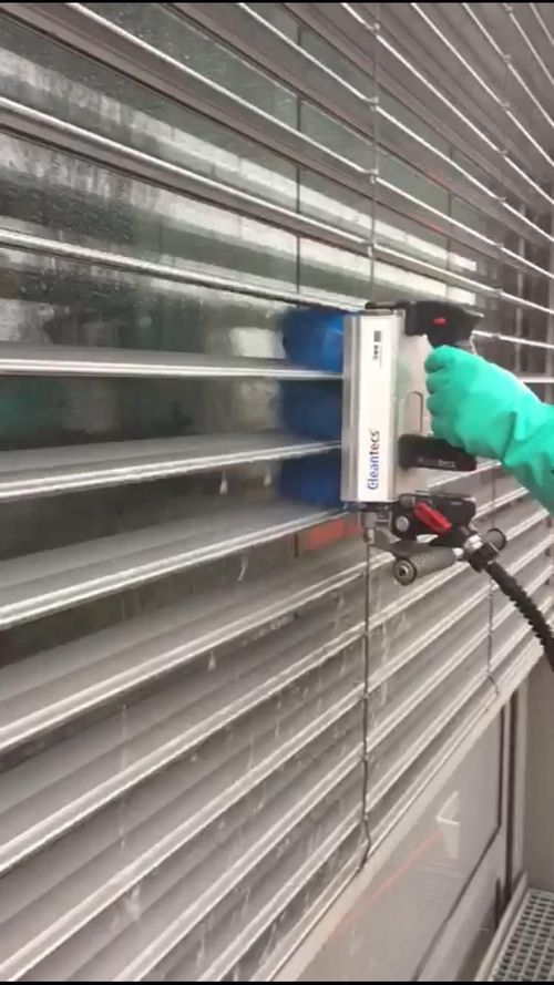 Eine Person mit schwarzen Handschuhen reinigt weisse Jalousien mit einem blauen Tuch und einer Sprühflasche.